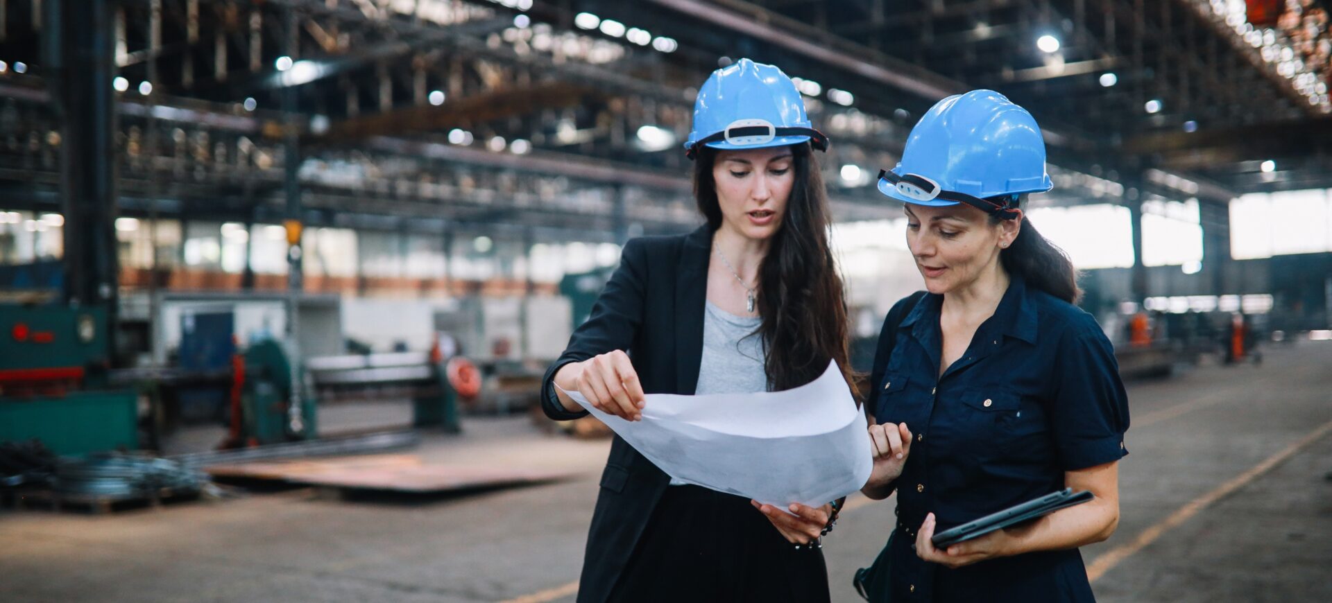 Blog 1 Two female engineers wearing blue hard hats examine blueprints in a factory setting.
