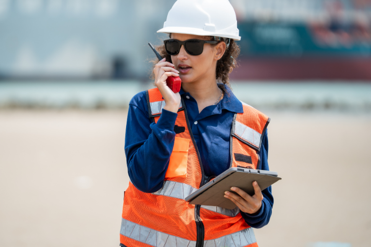 female port supervisor in a white hard hat, sunglasses, and orange safety vest holding a walkie-talkie and a tablet outdoors