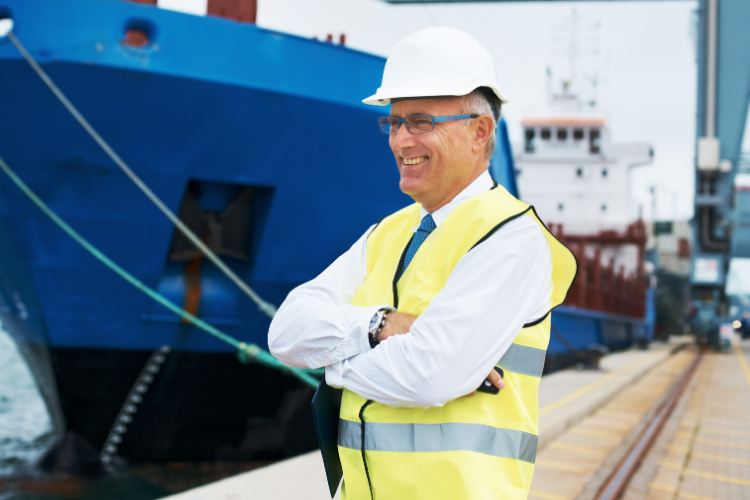 port manager in a white hard hat, tie, and high-visibility safety vest standing with arms crossed at the dockside with ships in the background