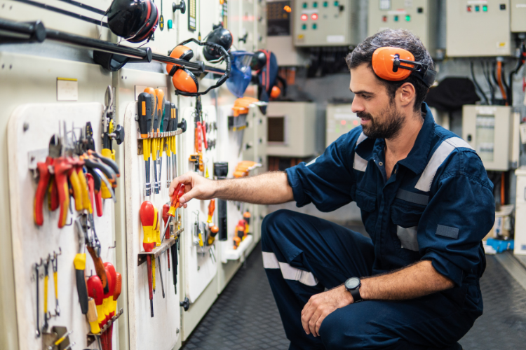 marine technician in blue coveralls and ear protection kneeling in a ship's engine room, selecting a tool from a wall-mounted tool board