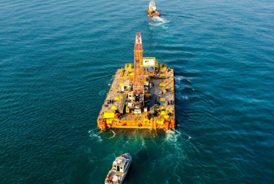 Aerial shot of heavy equipment on a barge in the sea, supported by tugboats.