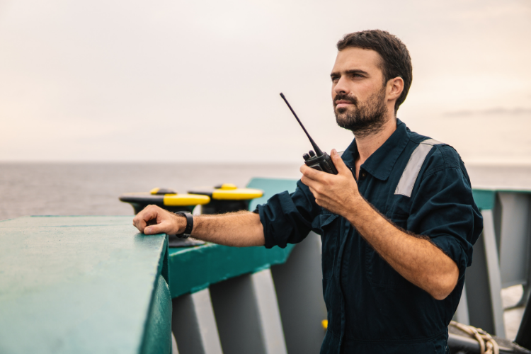 A engineer in a blue uniform using a walkie-talkie while standing on the deck of a ship