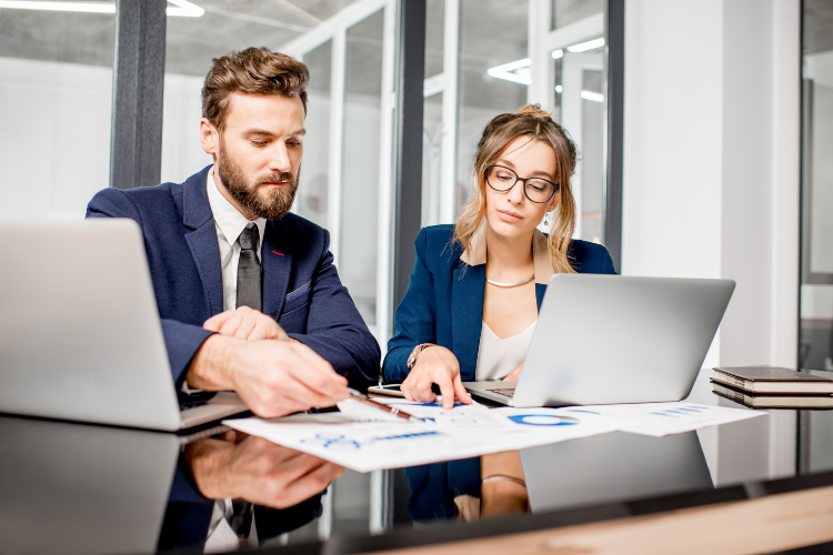 A businessman and businesswoman in professional attire reviewing charts and documents at an office desk with laptops