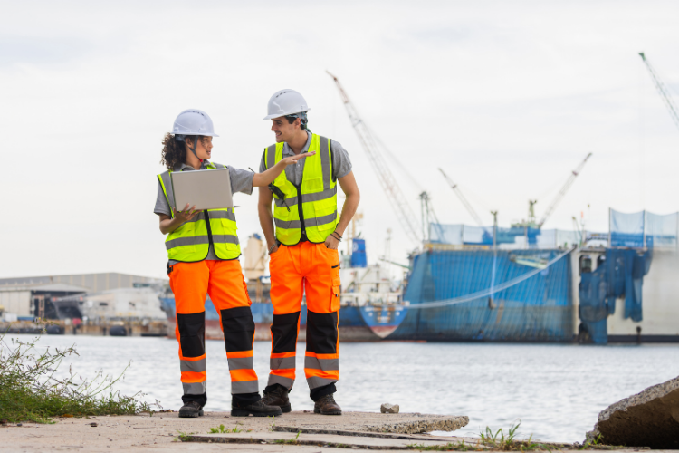 Two maritime engineers wearing high-visibility gear and hard hats reviewing data on a laptop by the shipyard