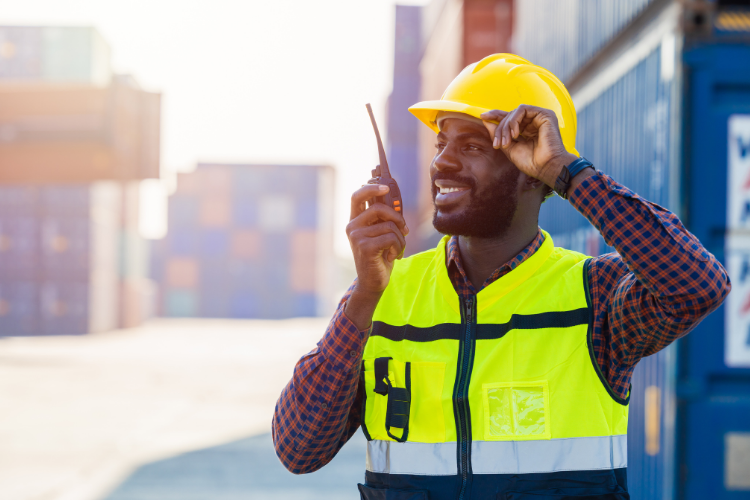 A port worker in a yellow hard hat and high-vis vest using a walkie-talkie outdoors among shipping containers