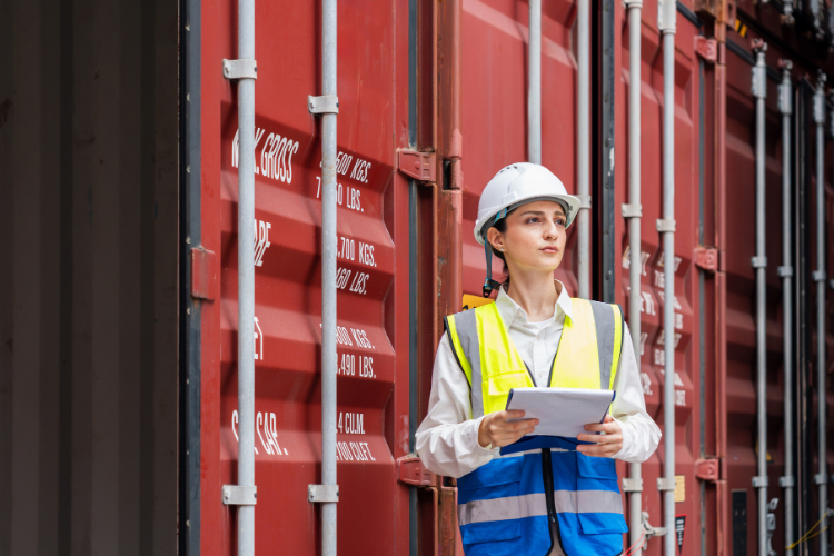 A female port logistics inspector wearing a hard hat and safety vest holding a clipboard in front of red shipping containers