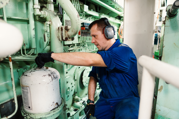 marine engineer wearing a blue uniform, gloves, and ear protection operating a valve on a large ship engine