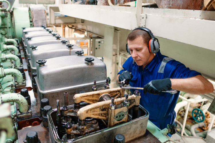 A marine engineer wearing a blue uniform and ear protection using a wrench to service a large ship engine