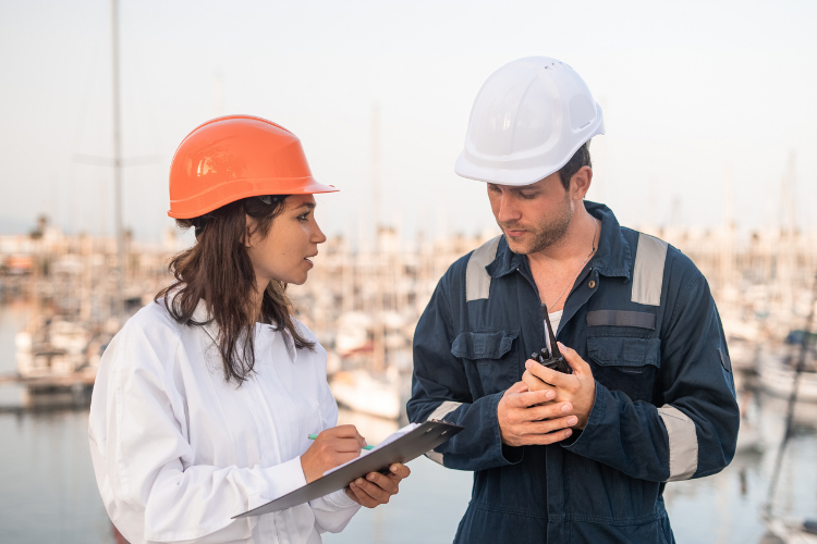 A female inspector in an orange hard hat writing on a clipboard while talking to a male marine worker holding a walkie-talkie at a marina