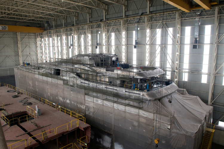 A large luxury yacht hull under construction inside a spacious industrial shipyard