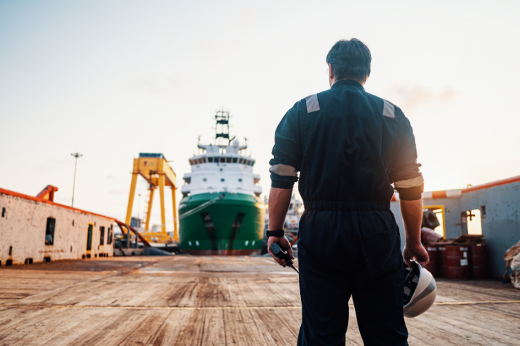 marine worker in a dark uniform holding a hard hat and device while looking out at a large ship docked at a shipyard