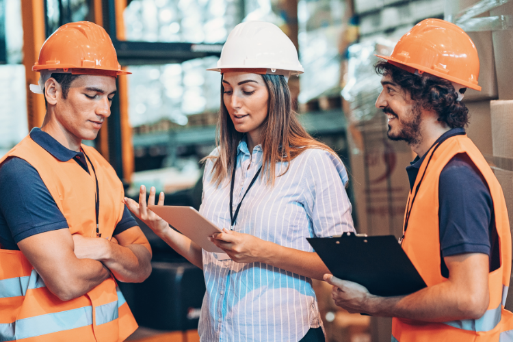 female supervisor in a white hard hat holding a tablet while discussing logistics with two workers wearing orange hard hats and safety vests