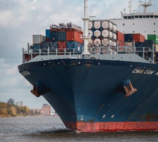 A large container ship moving through a river past a distant city shoreline